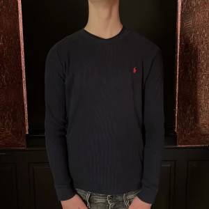 Ralph Lauren marinblå tröja - Säljer en stilren mörkblå tröja från Ralph Lauren med lång ärm och ribbad struktur. Den har en liten röd logotyp på bröstet. Perfekt för en avslappnad och snygg look. Säljer för en bra pris. Valentino