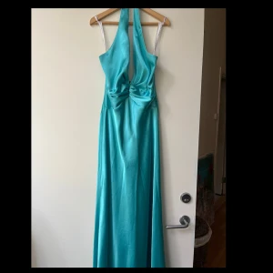 Turkos balklänning - Elegant turkos långklänning med öppen rygg och djup urringning. Klänningen har en vacker drapering framtill och är perfekt för speciella tillfällen. Den är ärmlös och har en smickrande passform.