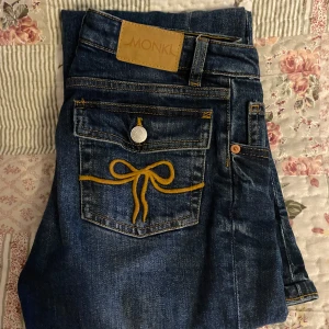 Blå jeans från Monki - Snygga blå jeans från Monki med unika broderade detaljer i form av gula rosetter på bakfickorna. De har en klassisk femficksdesign och knappgylf. Lågmidjade och bootcut. Pris kan diskuteras vid snabb affär!