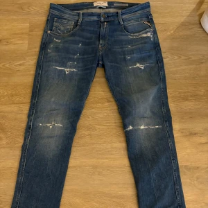 Blå jeans från Replay - Snygga blå jeans från Replay med slitna detaljer och en klassisk femficksdesign. De har en knappgylf och är perfekta för en avslappnad stil. Märkesloggan syns på insidan av fickan.