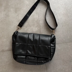 Svart quiltad axelväska - Snygg svart axelväska i quiltat mönster med justerbar axelrem. Väskan har en stilren design och stängs med magnetknappar. Perfekt för att bära dina dagliga nödvändigheter med stil.