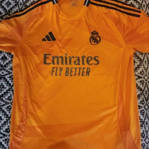 Orange fotbollströja från Adidas - Det är en reprint. Säljer en snygg orange fotbollströja från Adidas med Real Madrids emblem och sponsorlogga på framsidan. Tröjan har korta ärmar och tre svarta ränder på axlarna. Perfekt för fotbollsfans som vill visa sitt stöd. 🧡 storlek XL