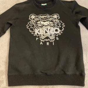 Svart sweatshirt från Kenzo - Säljer en svart sweatshirt från Kenzo med ett stort broderat tigerhuvud och texten 'KENZO PARIS' på framsidan. Tröjan har långa ärmar och en rund halsringning. Perfekt för en stilren och trendig look.