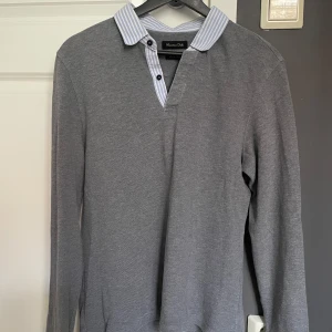 Grå tröja från Massimo Dutti - Säljer en stilren grå tröja från Massimo Dutti med en randig krage i blått och vitt. Tröjan har långa ärmar och en knappslå med två knappar. Perfekt för en avslappnad men ändå elegant look.