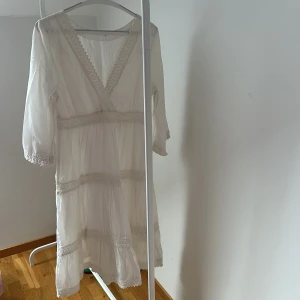 Vit klänning från Melmelli - Charmig vit klänning från Melmelli med v-ringad design och spetsdetaljer. Klänningen har trekvartsärmar och en luftig passform, perfekt för varmare dagar. Den romantiska stilen gör den till ett mångsidigt plagg i garderoben.