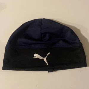 Svart och blå mössa från Puma - Snygg och sportig mössa från Puma i svart och blått. Mössan har en vit Puma-logga framtill och är perfekt för kyligare dagar. Den är tillverkad i ett mjukt och bekvämt material.