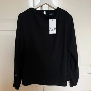 Svart blus från Zara - Elegant svart blus från Zara med långa ärmar och en dekorativ knappdetalj i nacken. Perfekt för en stilren look. Blusen har en enkel och tidlös design som passar till många olika tillfällen.