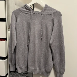 Grå hoodie - Säljer en stilren grå hoodie med dragsko i huvan. Tröjan har långa ärmar och ribbade muddar vid ärmslut och nederkant. Perfekt för en avslappnad look.