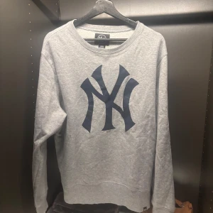 Grå sweatshirt med tryck från '47 - Säljer en grå sweatshirt från '47 med ett stort mörkblått tryck på framsidan. Tröjan är tillverkad i en bomulls- och polyesterblandning och har långa ärmar. Perfekt för en avslappnad stil.