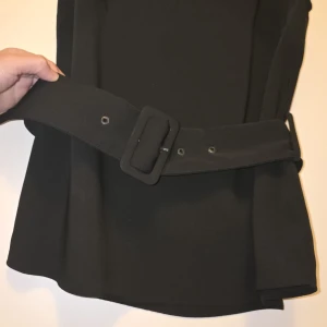 Svart bluz med bälte - Elegant svart klänning med ett matchande bälte i midjan. Klänningen har en stilren design och är ärmlös, vilket gör den perfekt för olika tillfällen. Den har en figurnära passform som framhäver silhuetten.