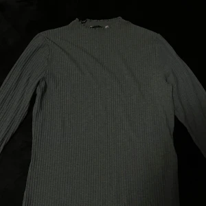 Grå/grön ribbad tröja - Säljer en stilren grå ribbad tröja med långa ärmar. Perfekt för en avslappnad och bekväm look. Tröjan har en hög hals och är tillverkad i ett mjukt material.