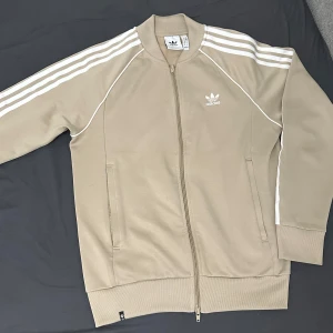 Beige kofta från Adidas - Snygg beige kofta från Adidas med klassiska vita ränder längs ärmarna. Jackan har dragkedja framtill och två fickor. Perfekt för en sportig look.