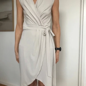 Beige klänning Stl xs Ny -  klänning  ljus kittbeige färg med en v-ringning och korta ärmar. H&M. Den har en knäppning i sidan, omlott knyt i midjan. Glipar inte, sitter bra på. mjuk, stretchig och bekväm kvalitet Poly/visc. Nyskick. Stl XS, passar en small bra.