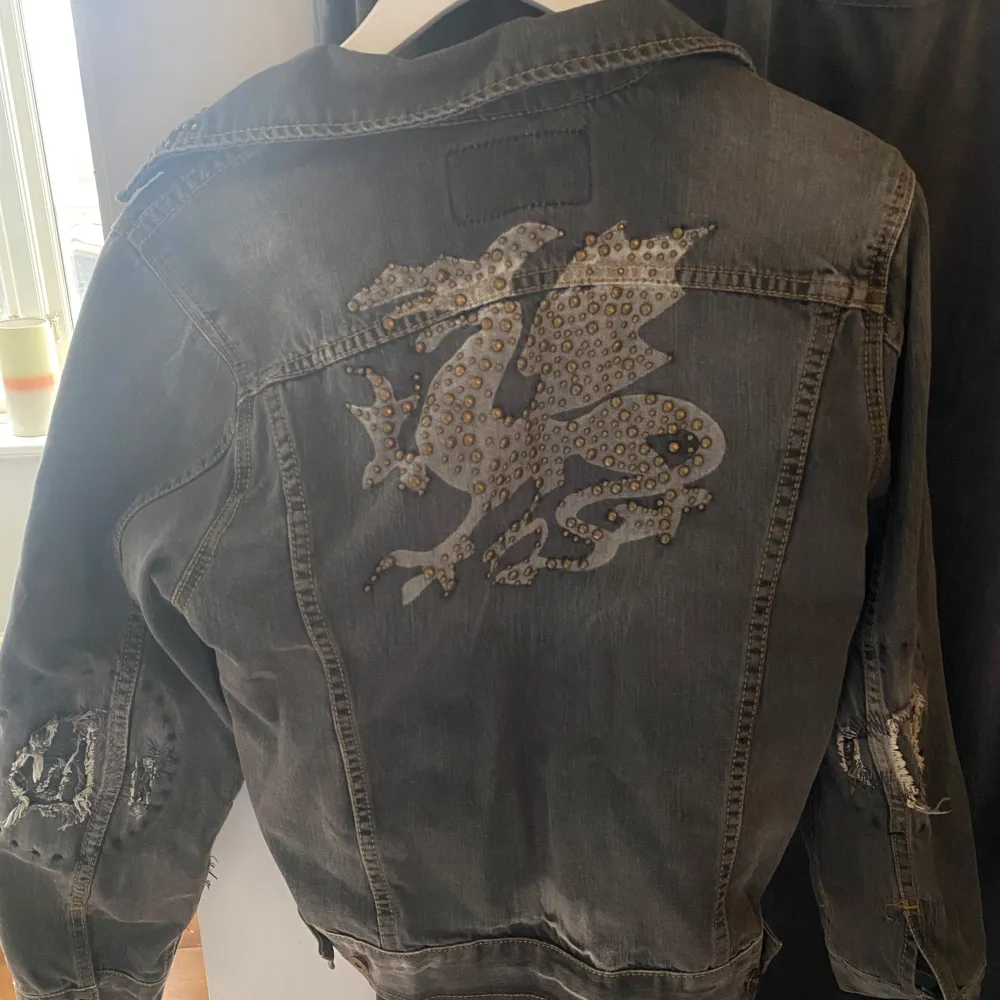 Cool svart jeansjacka från Replay med unika detaljer. Jackan har nitar på kragen och fickorna samt ett stort drakmönster på ryggen. Den har slitna detaljer på ärmarna och en broderad logga i orange. Köpt vintage i Paris💓. Takit.