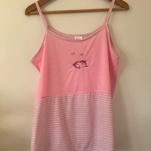 Rosa randig linnetopp med delfinbroderi - Söt rosa linnetopp med smala axelband och ett broderi av två delfiner framtill. Toppen har en randig nederdel i ljusrosa och vitt. Perfekt för en avslappnad sommarlook.