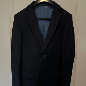 Dressman Mörkblå kavaj  - Elegant mörkblå kavaj med två knappar framtill och en stilren passform. Kavajen har en ljusblå insida med röda detaljer vid fickorna. Perfekt för formella tillfällen eller fester. Säljer pågrund av att jag inte har någon användning av den längre. Använd på skolavslutningen som sedan dess har legat i min garderob och plockat på damm. Om ni kommer med frågor så är det bara att höra av sig, är väldigt aktiv här på Plick.