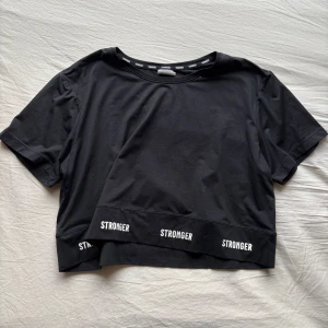 Svart t-shirt från Stronger - Snygg svart t-shirt från Stronger med textdetaljer längs fållen och halsen. Tillverkad av 90% återvunnen polyester och 10% elastan perfekt för en aktiv livsstil. Kortärmad design som ger en sportig look.