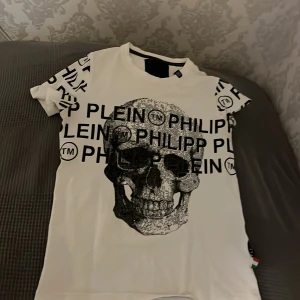 Vit t-shirt med dödskalle från Philipp Plein - Säljer en cool vit t-shirt från Philipp Plein med ett stort dödskalletryck på framsidan. T-shirten har korta ärmar och märkesnamnet tryckt flera gånger över hela plagget. Perfekt för dig som vill sticka ut med en unik stil.