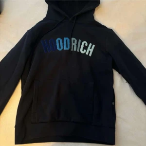 Svart hoodie från Hoodrich - Säljer en svart hoodie från Hoodrich med text i blå och vit nyans på bröstet. Den har en klassisk känguruficka och justerbar huva med dragsko. Perfekt för en avslappnad stil.
