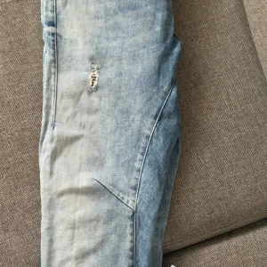 Ljusblå jeans med slitningar - Säljer ett par ljusblå jeans med fabriksgjorda slitningar på framsidan. De har en snygg passform och är perfekta för en avslappnad stil. Jeansen har klassiska fem fickor och en snygg tvättad look.