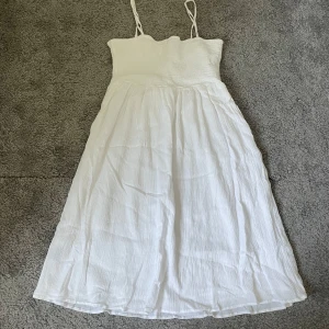 Vit sommarklänning med smala axelband - Säljer en superfin vit sommarklänning med smala axelband. Klänningen har en smockad överdel som ger en bekväm passform och ett flödande kjolparti som är perfekt för varma dagar. Den är gjord i ett lätt och luftigt material, vilket gör den idealisk för sommarens alla äventyr. Perfekt för både stranden och sommarfester!