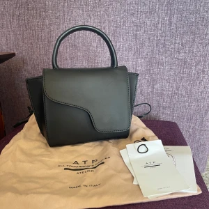 ATP atelier miniväska  - Säljer denna fina ATP-atelierväska i svart. Väskan köptes ny på boozt.com för 3 900 kr men då den är något repig (se bilder) säljs den för 2500 kr. Dustbag och skötselråd skickas med.Väskan är perfekt till bal/finsitrning tex och man kan ta av banden