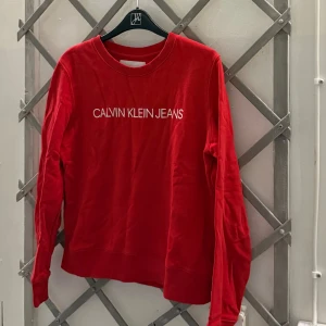 Tröja calvin klein - En röd tröja med en vit Calvin Klein Jeans-logotyp på framsidan. Tröjan har en rund halsringning och långa ärmar. Den är använd fåtal gånger och är i väldigt fint sick.