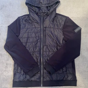 Svart jacka från Barbour - Säljer en snygg svart jacka från Barbour i storlek XL men passar även L. Den har en quiltad design med dragkedja och en huva för extra skydd. Perfekt för höst och vår. Märkeslogga på ärmen ger en extra touch av stil. Passar både til