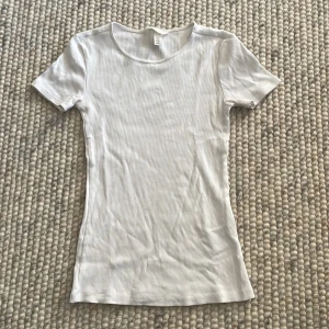 Vit ribbad t-shirt - Säljer en enkel och stilren vit ribbad t-shirt. Perfekt basplagg för alla tillfällen. Den har en tight passform och korta ärmar, vilket gör den idealisk för både vardag och fest. Materialet är mjukt och bekvämt.