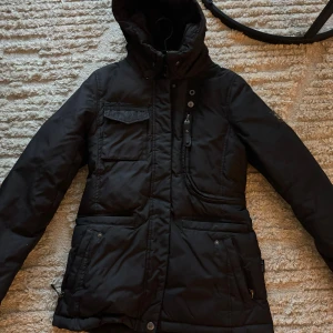 Svart vinterjacka från Hollies - Säljer en varm svart vinterjacka från Hollies. Jackan har en stilren design med flera praktiska fickor och en hög krage för extra skydd mot kylan. Den är perfekt för kalla vinterdagar och har en dragkedja samt knappar