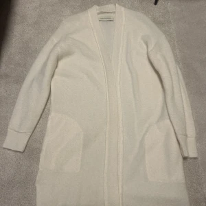 Vit kofta från By Malene Birger - Säljer en elegant vit kofta från By Malene Birger. Den är lång och har långa ärmar med ribbade muddar. Perfekt för att hålla sig varm och stilren under kyligare dagar. Koftan har en öppen design utan knappar, fickor fram och korta sprunn i sidan.