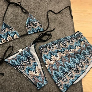 oanvänt mönstrat bikini set med kjol - Snygg bikini med ett färgglatt mönster i blått, svart och beige. Toppen har knytband och trekantsform, medan underdelen är en kjol med matchande mönster. Aldrig använd!
