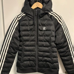 Svart pufferjacka från Adidas - Snygg svart pufferjacka från Adidas med klassiska vita ränder längs ärmarna. Jackan har en huva och dragkedja framtill. Perfekt för kyligare dagar med sin vadderade design.