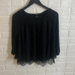 Svart blus från Lindex - Elegant svart blus från Lindex med långa ärmar och vackra spetsdetaljer vid nederkanten. Blusen har ett plisserat mönster framtill som ger en stilren look. Perfekt för en chic och sofistikerad stil.
