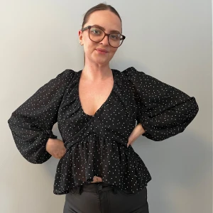 Svart prickig blus med öppen rygg - Snygg svart blus med vita prickar och en v-ringad design. Blusen har långa ärmar och en öppen rygg med knytband. Perfekt för en stilren look.