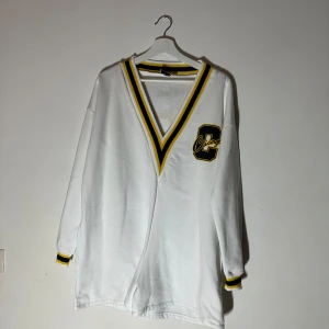 Vit långärmad tröja från ASOS DESIGN - Säljer en vit långärmad tröja från ASOS DESIGN med ett stort 'Chicago' tryck på ryggen och en gul och svart randig v-ringning. Perfekt för en avslappnad stil med en sportig touch.🥰🥰🥰