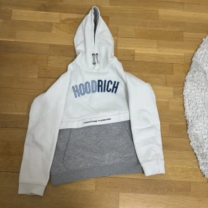 Vit och grå hoodie från Hoodrich - Säljer en snygg vit och grå hoodie från Hoodrich med texten 'HOODRICH' i blått på bröstet. Hoodien har en stor ficka framtill och en justerbar huva. Perfekt för en avslappnad stil. Passar åldrar 13-15 år.