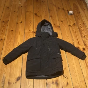 Svart vinterjacka från Didriksons - Säljer en svart vinterjacka från Didriksons, perfekt för kalla och blöta dagar. Jackan är 100% vattentät och har en huva för extra skydd. Den har långa ärmar och praktiska fickor framtill. Insidan är fodrad för extra värme.