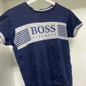 Blå t-shirt från Hugo Boss - Snygg blå t-shirt från Hugo Boss med grått tryck och texten 'BOSS' på bröstet. Den har korta ärmar med gråa kanter och en rund halsringning. Perfekt för en avslappnad stil.