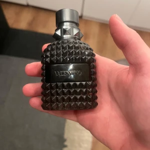 Valentino Parfym - Ungefär 40 ml kvar! Säljer en elegant parfym från Valentino med en stilren flaska i mörkt glas och nitar på korken. Flaskan har en lyxig och modern design som passar perfekt för den som vill ha något unikt.