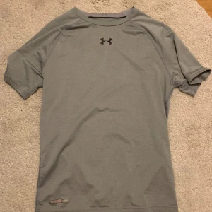 Grå t-shirt från Under Armour - Säljer en grå t-shirt från Under Armour med korta ärmar och rund hals. Perfekt för träning med sitt funktionella material. Diskret logga på bröstet och nacken. Slim