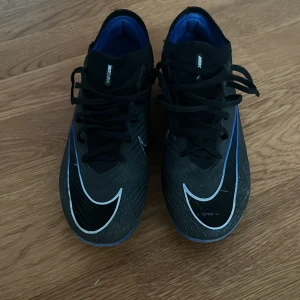 Svarta Nike Elite Mercurial fotbollsskor - Säljer ett par svarta Nike Mercurial fotbollsskor med vita och blå detaljer. Skorna säljs pågrund av för små, har ungefär använts i 3 månader men inte så använda pågrund av skada. Köpte för 2800 kr. Pris kan diskuteras.