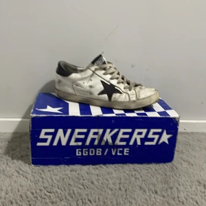Golden Goose Sneakers Superstar - Golden Goose Sneakers Superstar i vitt med svarta detaljer och en krämfärgad sula. Skorna har en ikonisk stjärndetalj på sidan och snörning framtill. Tillverkade i Italien, dessa sneakers kombinerar stil och komfort.