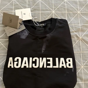 Svart t-shirt från Balenciaga storlek XL men sitter som L - Säljer en svart t-shirt från Balenciaga med vit text på framsidan. T-shirten har en klassisk rund hals och korta ärmar. Perfekt för en stilren look.