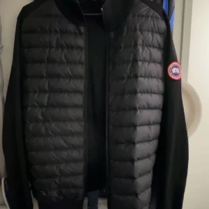 Svart pufferjacka från Canada Goose - Säljer en svart pufferjacka från Canada Goose med deras ikoniska märke på ärmen. Jackan har en quiltad design och dragkedja framtill. Perfekt för kyligare väder och en stilren look. Den är äkta köpt för 7200 på johnells. 