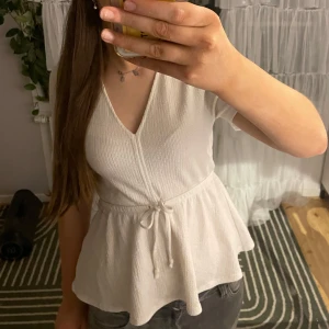 Vit ribbad blus med knytband - Säljer en stilren vit blus med v-ringning och knytband i midjan. Blusen har korta ärmar och en ribbad struktur som ger en fin textur. Perfekt för en avslappnad men ändå elegant look.
