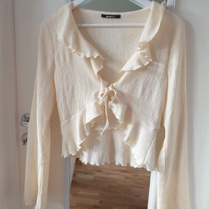 Beige blus från Gina Tricot - Säljer en söt beige blus från Gina Tricot med volangdetaljer och knytning framtill. Blusen har långa ärmar och en lätt textur i tyget. Perfekt för en stilren look!