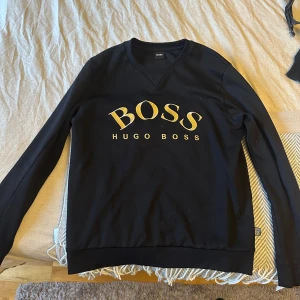 Svart tröja från Hugo Boss - Säljer en stilren svart tröja från Hugo Boss med guldtryck på framsidan. Tröjan har långa ärmar och en rund halsringning.