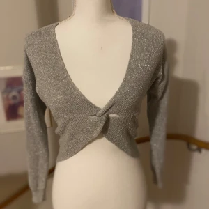 Glittrig grå/silvrig kofta från Sisley - Snygg glittrig grå/silvrig kofta från Sisley med en knutdetalj framtill. Perfekt för att lägga till lite glamour till din outfit. Koftan har långa ärmar och en djup v-ringning.