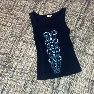 Svart linne med blått mönster - Snygg svart linne med ett unikt blått mönster framtill. Perfekt för att ge en cool touch till din outfit. Toppen har en rund halsringning och är tillverkad i ett mjukt material.